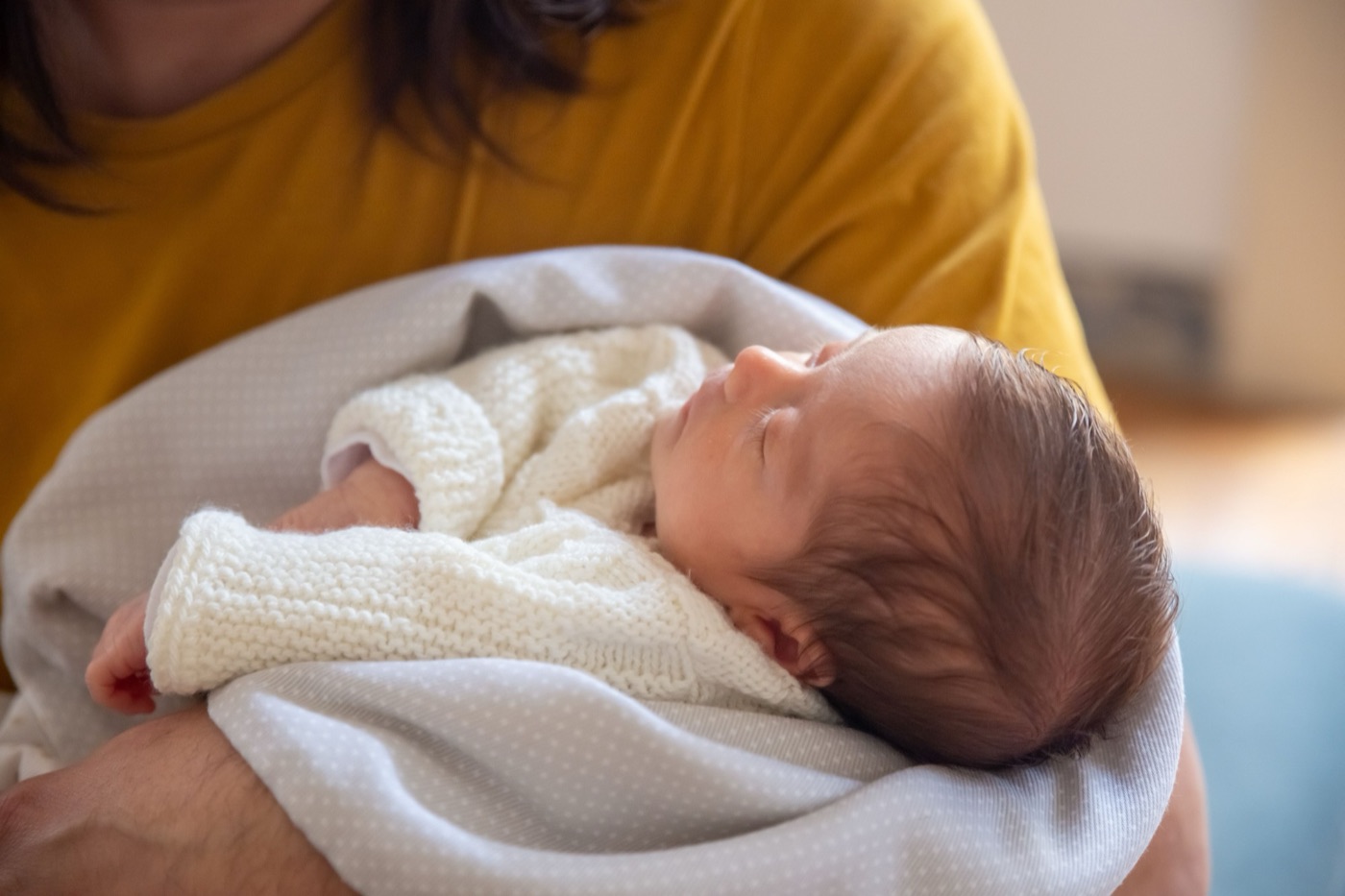 Noworodek w wełnianym sweterku otulony kocykiem śpi na rękach mamy