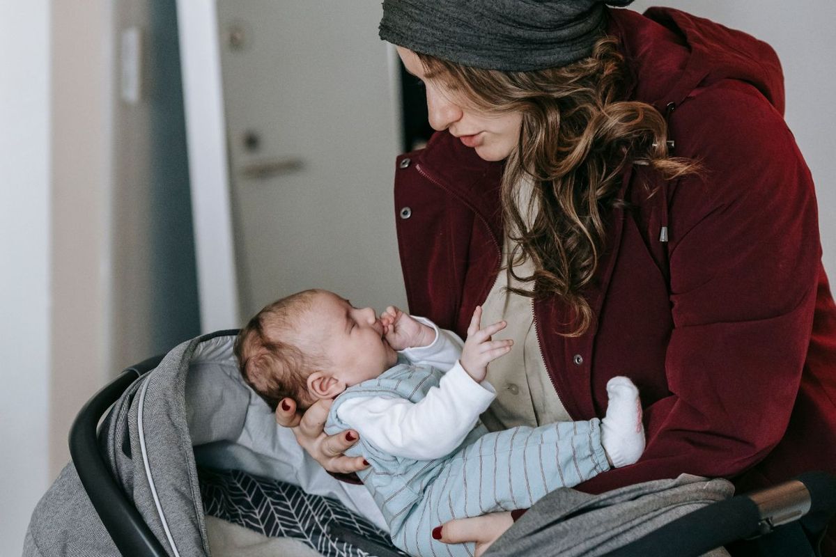 Mama w kurtce wkłada noworodka w cienkim pajacyku do fotelika samochodowego